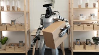 A silver humanoid robot with a black head module holding a cardboard box against its chest in front of wooden shelves