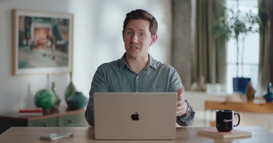 A man in a grey button-up shirt speaking to the camera behind an open silver laptop at a wooden desk, with a black OpenAI mug, green vases and a framed painting in a bright room