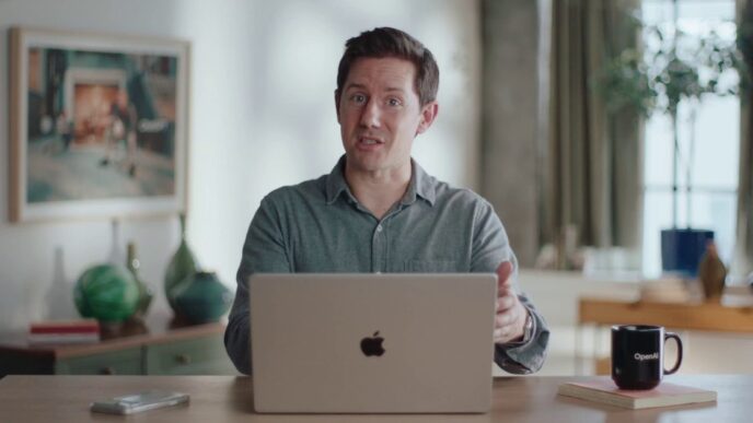 A man in a grey button-up shirt speaking to the camera behind an open silver laptop at a wooden desk, with a black OpenAI mug, green vases and a framed painting in a bright room