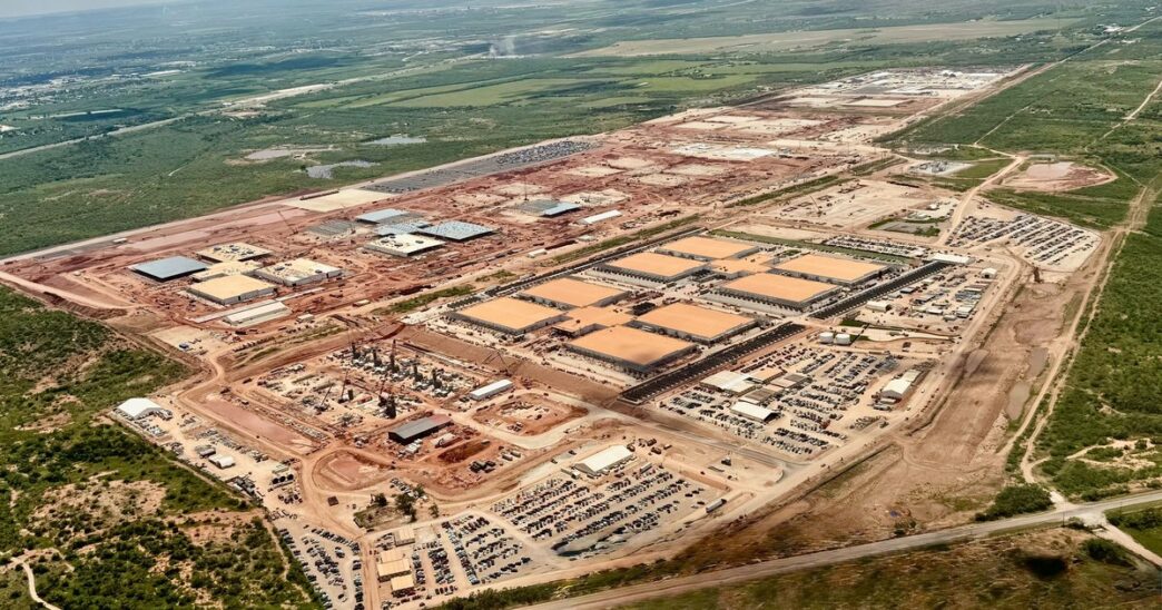 Aerial view of OpenAI's Stargate I data center site in Abilene, Texas, under construction, with rows of tan-roofed buildings, parking lots and red earth surrounded by green Texas scrubland