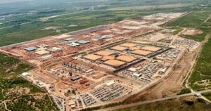 Aerial view of OpenAI's Stargate I data center site in Abilene, Texas, under construction, with rows of tan-roofed buildings, parking lots and red earth surrounded by green Texas scrubland