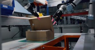 Two silver collaborative robot arms with pink-tipped grippers opening a cardboard return parcel on a packing station in a warehouse