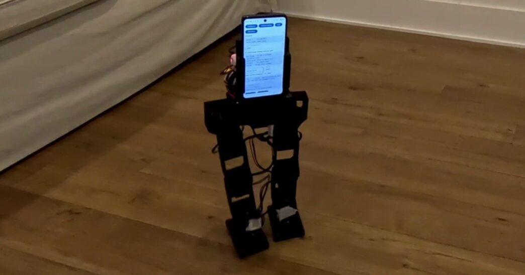 The small black PhoneBot biped robot walks on a wood floor beside a sofa, with an Android phone mounted as its torso