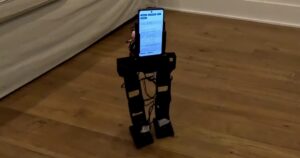 The small black PhoneBot biped robot walks on a wood floor beside a sofa, with an Android phone mounted as its torso