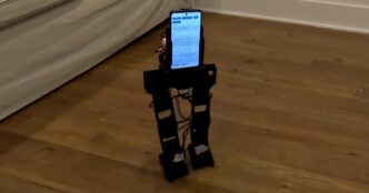 The small black PhoneBot biped robot walks on a wood floor beside a sofa, with an Android phone mounted as its torso