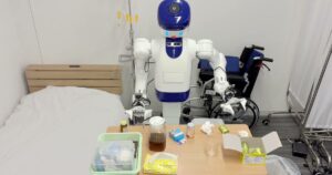 A white and blue robot with two arms and a round camera head working at a bedside table with a pitcher, tissues and medicine bottles next to a hospital bed and a wheelchair