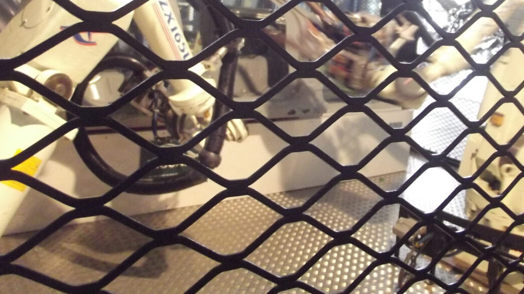 White industrial robot arms seen through a black diamond-mesh safety fence, standing on a metal diamond-plate floor