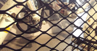 White industrial robot arms seen through a black diamond-mesh safety fence, standing on a metal diamond-plate floor