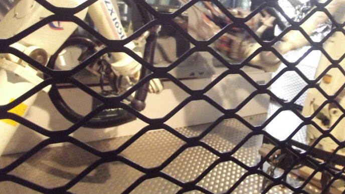 White industrial robot arms seen through a black diamond-mesh safety fence, standing on a metal diamond-plate floor