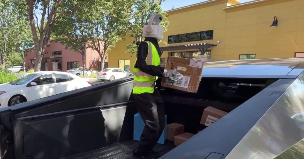 A humanoid robot with a white camera head, black jacket and yellow high-visibility vest standing in the bed of a Tesla Cybertruck holding a cardboard delivery box, with more boxes at its feet and shops behind