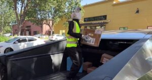 A humanoid robot with a white camera head, black jacket and yellow high-visibility vest standing in the bed of a Tesla Cybertruck holding a cardboard delivery box, with more boxes at its feet and shops behind