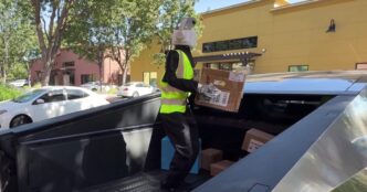 A humanoid robot with a white camera head, black jacket and yellow high-visibility vest standing in the bed of a Tesla Cybertruck holding a cardboard delivery box, with more boxes at its feet and shops behind