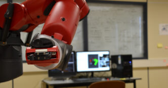 Close-up of a Rethink Robotics Baxter robot arm and its parallel-jaw gripper holding a small circuit board in a lab
