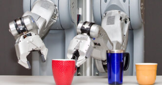 Two arms of a Willow Garage PR2 robot with grippers positioned over a red mug, a blue travel mug and a yellow cup on a table against a dark grey background