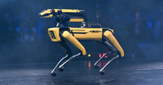 Yellow and black Boston Dynamics Spot quadruped robot standing on a dark stage under blue light