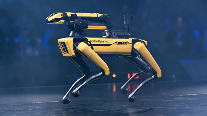 Yellow and black Boston Dynamics Spot quadruped robot standing on a dark stage under blue light
