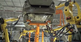Yellow FANUC industrial robot arms with cable bundles working around a car body held on an orange fixture in an automotive plant