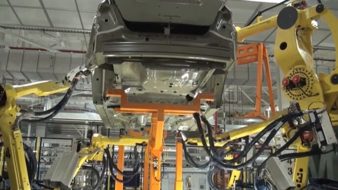 Yellow FANUC industrial robot arms with cable bundles working around a car body held on an orange fixture in an automotive plant