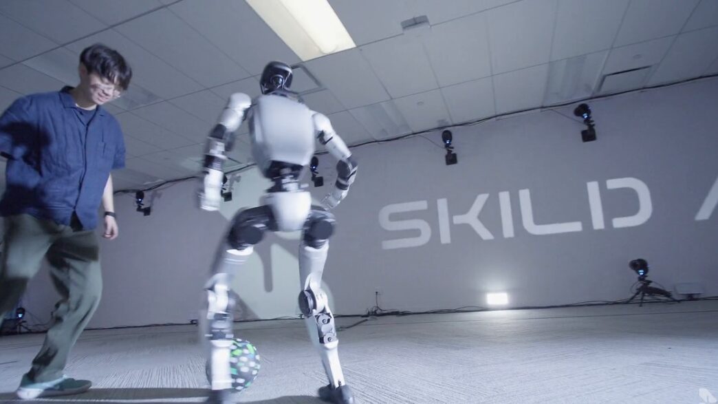 A white bipedal humanoid robot controls a soccer ball while a man defends, in front of a Skild AI wall logo