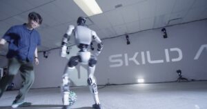 A white bipedal humanoid robot controls a soccer ball while a man defends, in front of a Skild AI wall logo