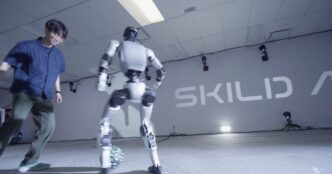 A white bipedal humanoid robot controls a soccer ball while a man defends, in front of a Skild AI wall logo
