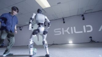 A white bipedal humanoid robot controls a soccer ball while a man defends, in front of a Skild AI wall logo