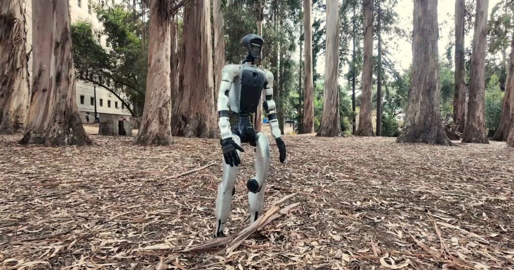 White Unitree G1 humanoid robot standing on a carpet of dry eucalyptus leaves among tall eucalyptus trees, from the SuperMimic project video
