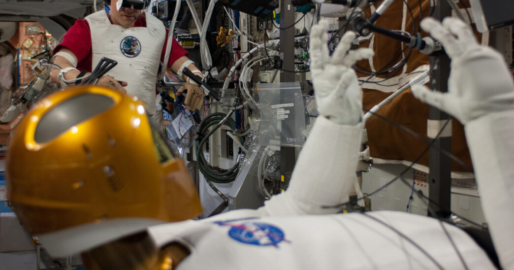 NASA astronaut Tom Marshburn wearing a VR headset and motion-tracking sensors on his arms in the background while the gold-helmeted Robonaut 2 humanoid robot in the foreground raises its gloved hands, aboard the International Space Station