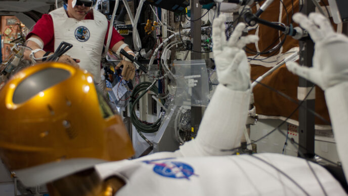 NASA astronaut Tom Marshburn wearing a VR headset and motion-tracking sensors on his arms in the background while the gold-helmeted Robonaut 2 humanoid robot in the foreground raises its gloved hands, aboard the International Space Station