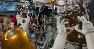 NASA astronaut Tom Marshburn wearing a VR headset and motion-tracking sensors on his arms in the background while the gold-helmeted Robonaut 2 humanoid robot in the foreground raises its gloved hands, aboard the International Space Station