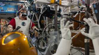 NASA astronaut Tom Marshburn wearing a VR headset and motion-tracking sensors on his arms in the background while the gold-helmeted Robonaut 2 humanoid robot in the foreground raises its gloved hands, aboard the International Space Station