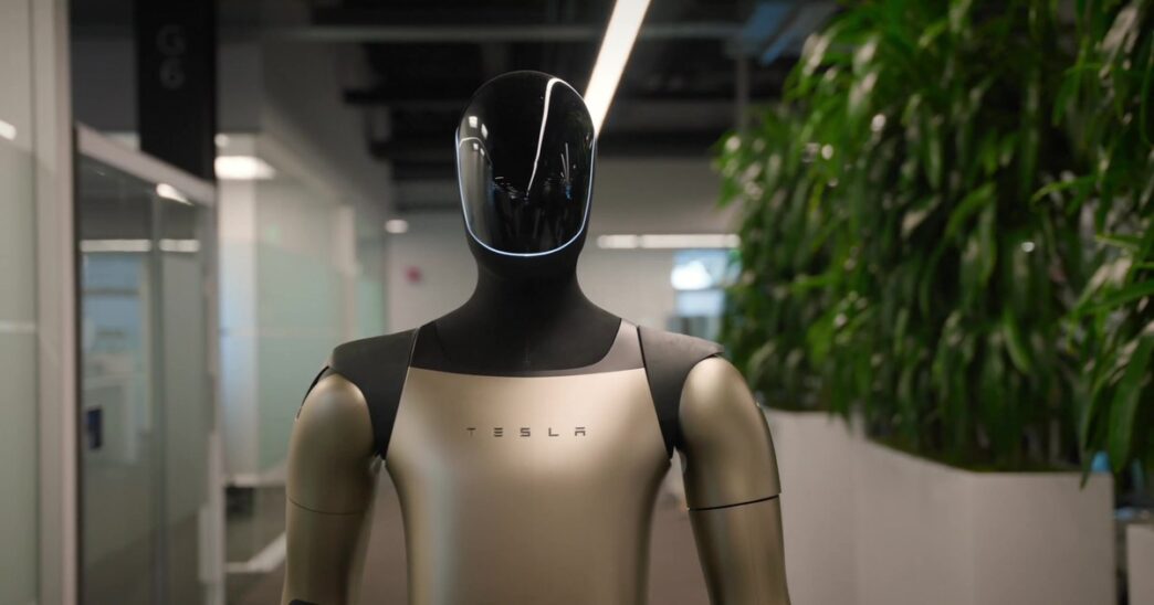 Tesla's Optimus humanoid robot with a black glossy head and champagne-colored torso marked TESLA, standing in an office corridor beside tall plants