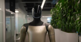 Tesla's Optimus humanoid robot with a black glossy head and champagne-colored torso marked TESLA, standing in an office corridor beside tall plants