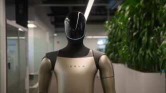 Tesla's Optimus humanoid robot with a black glossy head and champagne-colored torso marked TESLA, standing in an office corridor beside tall plants