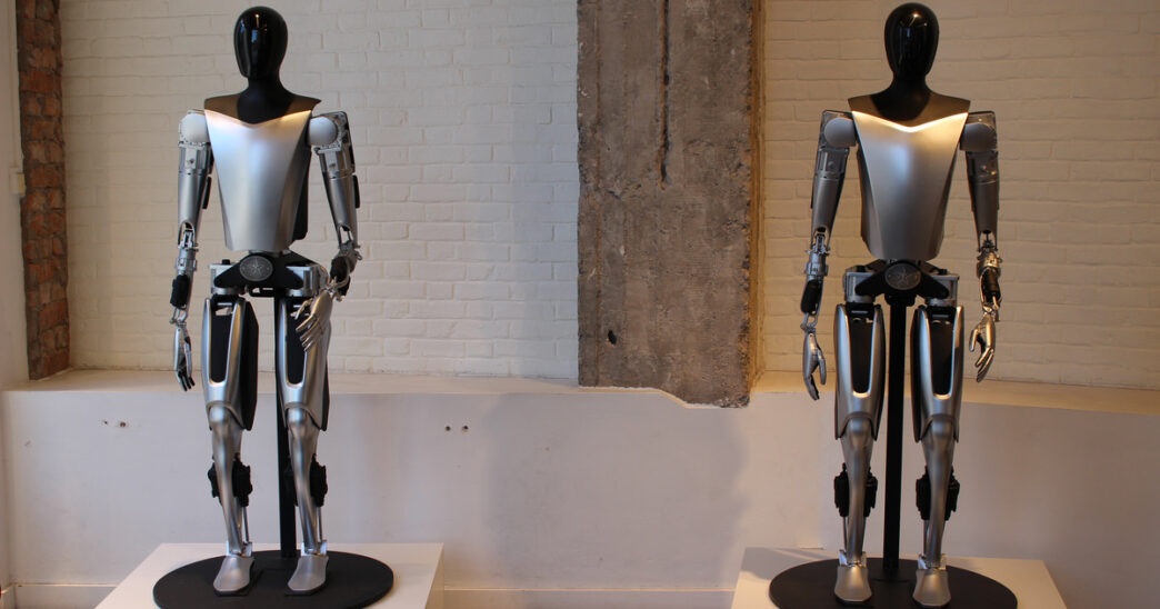 Two silver-and-black Tesla Optimus humanoid robot prototypes standing on pedestals in front of a white brick wall