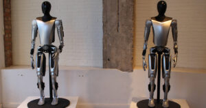 Two silver-and-black Tesla Optimus humanoid robot prototypes standing on pedestals in front of a white brick wall