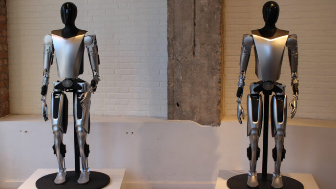 Two silver-and-black Tesla Optimus humanoid robot prototypes standing on pedestals in front of a white brick wall