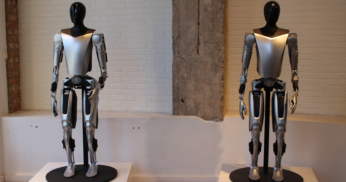 Two silver-and-black Tesla Optimus humanoid robot prototypes standing on pedestals in front of a white brick wall