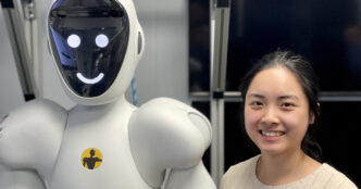 Smiling engineer standing beside a white humanoid robot whose face screen shows a simple smiley face, in front of an aluminium frame in a lab