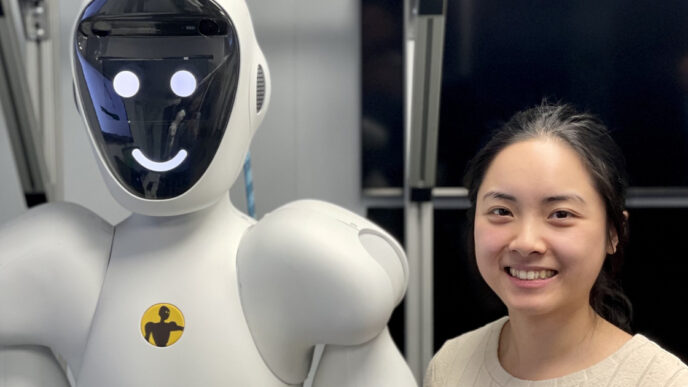 Smiling engineer standing beside a white humanoid robot whose face screen shows a simple smiley face, in front of an aluminium frame in a lab