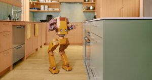 Tuesday Labs' orange two-legged Voxel robot with a boxy head, pixel eyes and two jointed arms standing in a wood-paneled home kitchen