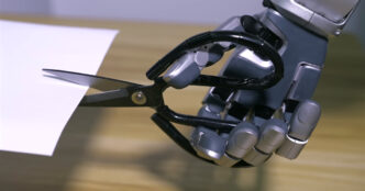 Grey and silver robotic hand gripping black scissors that are cutting a sheet of white paper on a wooden table