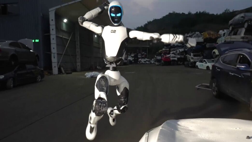 Unitree's white H2-X humanoid robot with a dark visor and glowing blue eyes leaping in mid-air with fists raised in a scrapyard at dusk