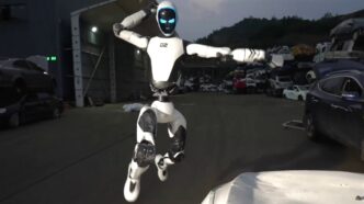 Unitree's white H2-X humanoid robot with a dark visor and glowing blue eyes leaping in mid-air with fists raised in a scrapyard at dusk