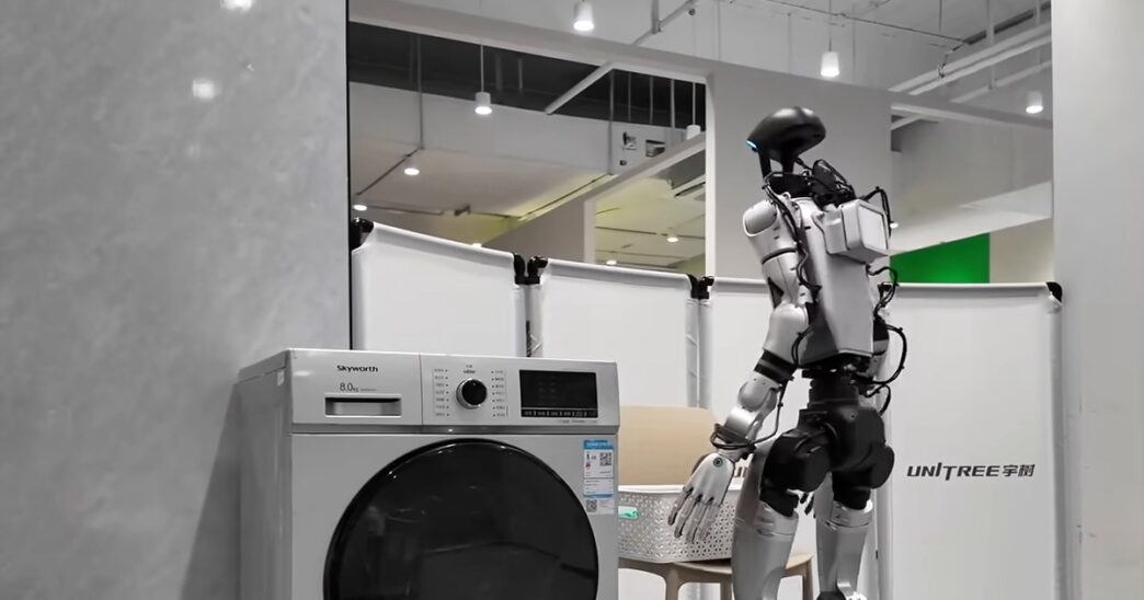 A silver and black Unitree humanoid robot with a glowing blue visor standing beside a front-loading washing machine and a laundry basket in a white test room with the Unitree logo on a partition