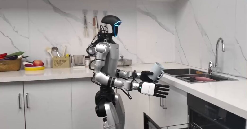 Silver and black Unitree humanoid robot standing in a white kitchen, holding a mug above an open dishwasher drawer