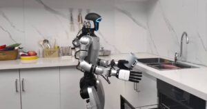 Silver and black Unitree humanoid robot standing in a white kitchen, holding a mug above an open dishwasher drawer