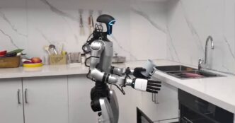 Silver and black Unitree humanoid robot standing in a white kitchen, holding a mug above an open dishwasher drawer