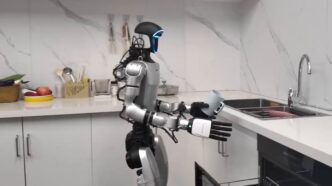 Silver and black Unitree humanoid robot standing in a white kitchen, holding a mug above an open dishwasher drawer