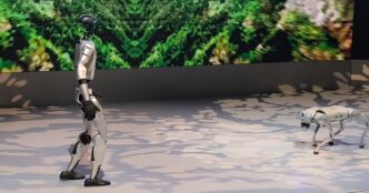 Unitree G1 humanoid robot walking across a stage at Japan Mobility Show 2025 with a robot dog in the background